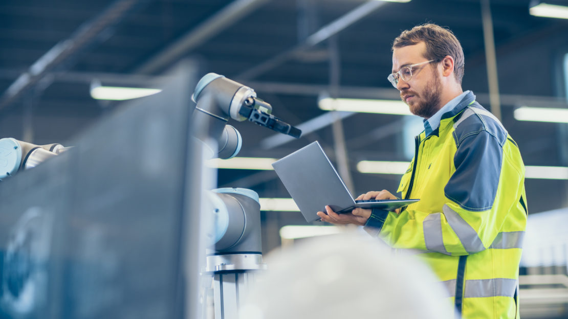 Factory worker programming a robotic arm using a laptop