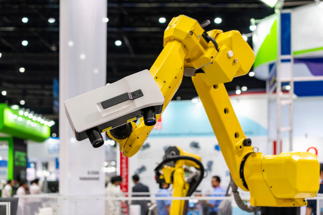 Photo of a yellow industrial robot with a camera/sensor attached to it. The robot is scanning an object during an expo.